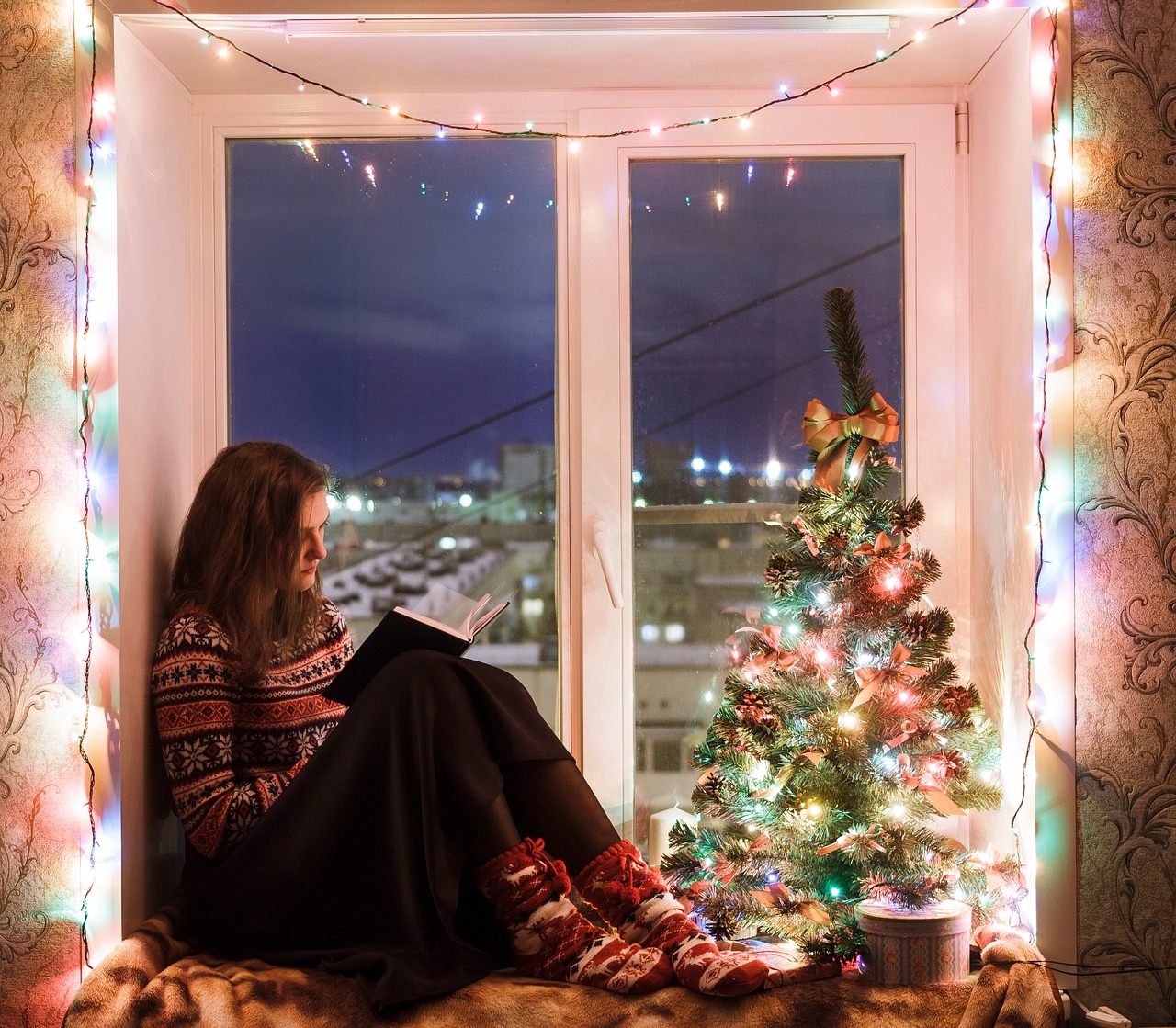 Woman sitting in window reading. Window decorated with Christmas lights.