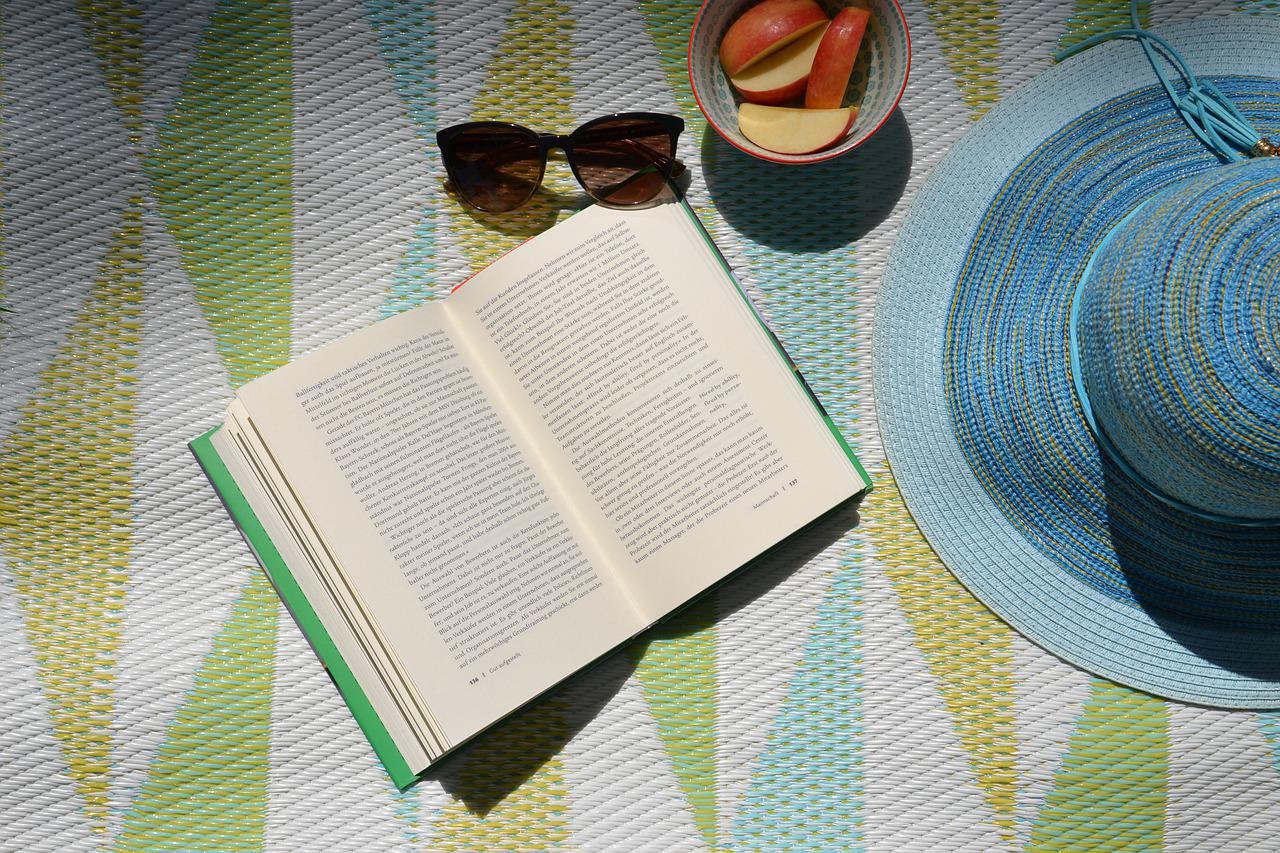 Blanket with book, sunhat, sunglasses, and apple slices sitting on it.