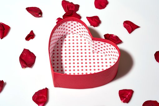 An empty heart shaped box with rose petals scattered around it.