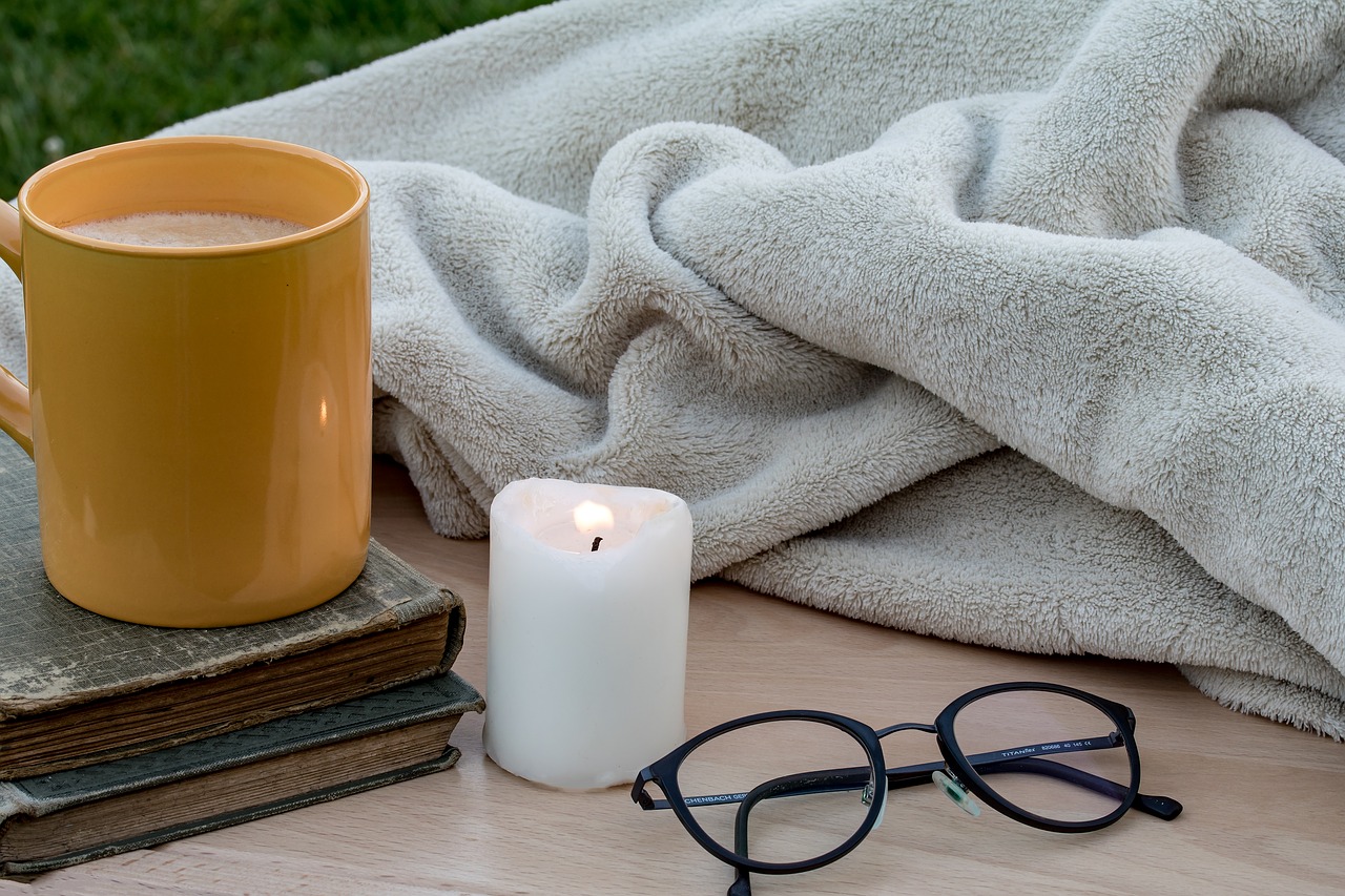 Close up of a candle, reading glasses, a blanket and a mug on two books.